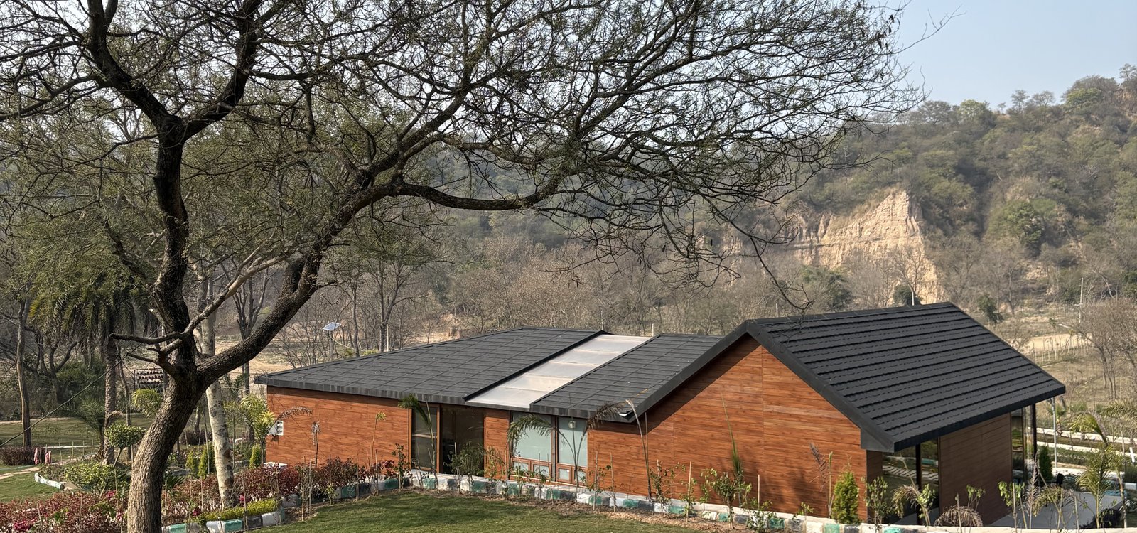 Eco-friendly farmhouse with a black roof, wooden exterior, large windows, and lush greenery against a scenic mountain backdrop.